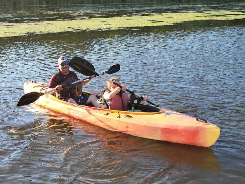 t: An adult and a young person paddle a tandem kayak together on a calm lake, the young person using an adaptive front seat, both enjoying the ride.