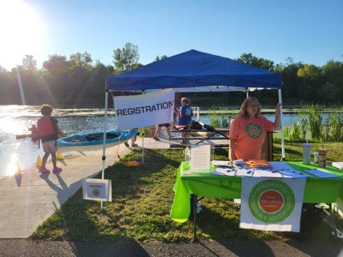 A golden hour beside the lake, a Rochester Accessible Adventures staff in an orange "Make Inclusion Happen" t-shirt welcomes guests at a green-draped registration table under a blue pop-up tent, with a kayak staged on the launch ramp behind her.