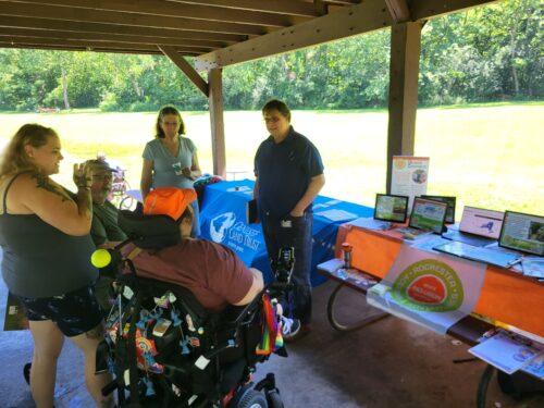A group of people gathered in the shade of a park pavilion, talking beside information tables for Rochester Accessible Adventures and Genesee Land Trust, with a grassy field and trees behind them.