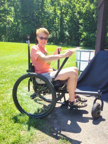 A smiling person in a coral Rochester Accessible Adventures t-shirt sits in a GRIT Freedom Chair — an all-terrain manual wheelchair with a large knobby front wheel and mountain-bike tires — on a park path in the sunshine.