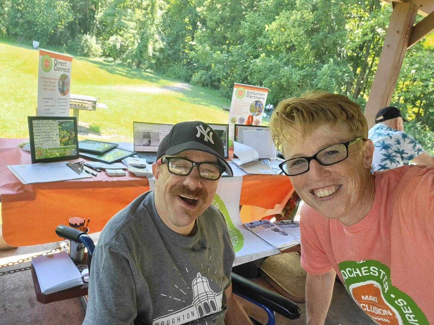 Two smiling people lean in for a selfie under the shade of a pavillion with an info table and a green grassy area and trees in the background