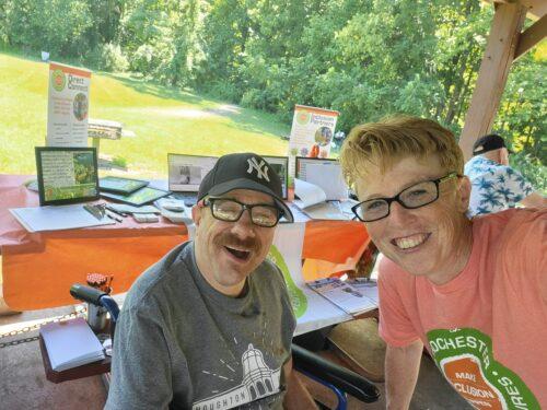 Two smiling people lean in for a selfie under the shade of a pavillion with an info table and a green grassy area and trees in the background