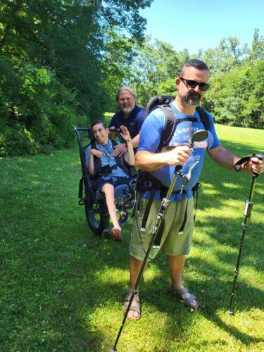 A man wearing a back harness is attached to his son who is in a Huckleberry Hiker chair; a man behind them supports the hiking chair