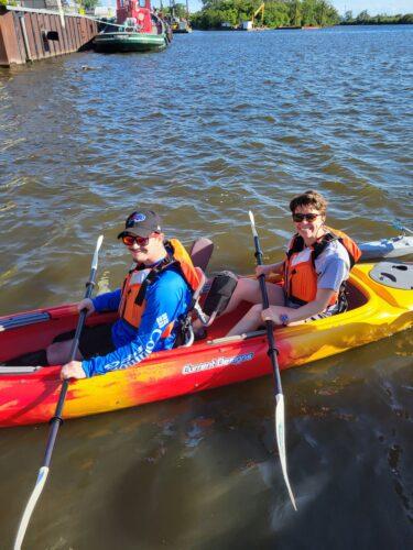 Two people smile up from a red and yellow tandem kayak in the water