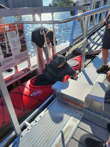A paddler situates himself in a red kayak on a Boardsafe Kayak launch at Elevator Alley with a staff person assisting from one side.