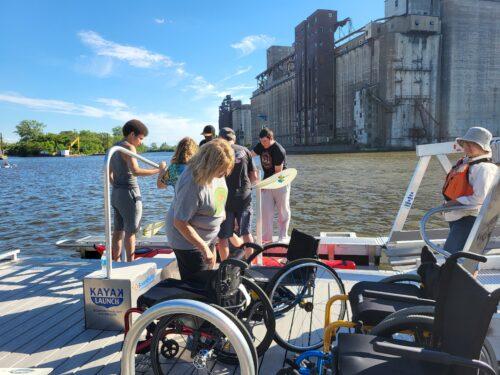 Dee and Elevator Alley Kayak staff assist guests into adaptive kayaks on a BoardSafe Kayak Launch with the grain elevators along a blue river in the background.