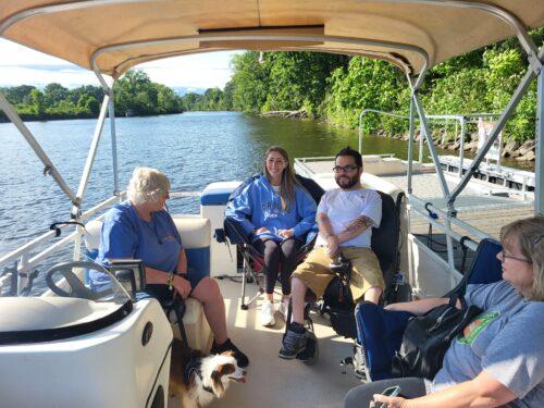 Four people get settled on a pontoon boat, each with their own seats, including one person in a power wheelchair.