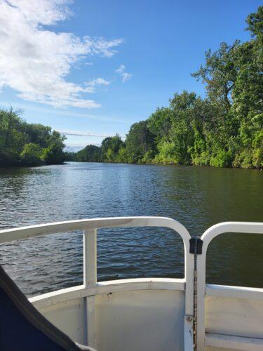 Railings in the foreground of a pontoon boat give way to a view of a wide canal lined by summer green trees under a brilliant blue sky.