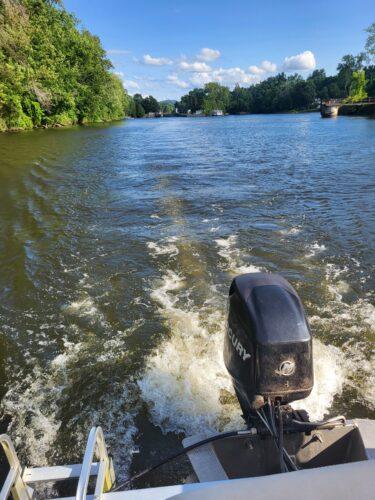 The view from behind looking out over the boat's motor shows a sun dappled forest of trees on one side and a shaded tree area on the right side with a wide expanse of canal between them.