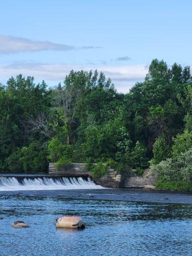 A calm river flows gently over a small dam, surrounded by dense green trees under a partly cloudy sky.