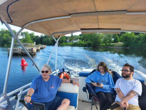 Four people on a pontoon boat with a shade overhead are cruising a wide canal, leaving a small wake behind them