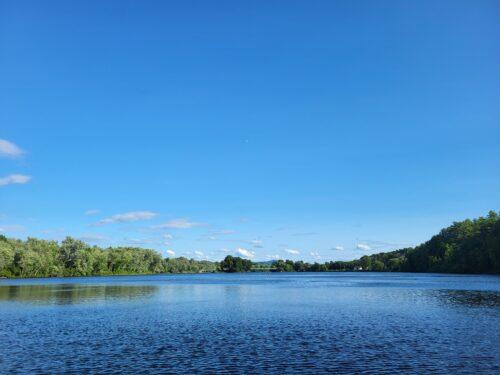 Big blue sky over a wide expanse of a river lined by summer green trees.