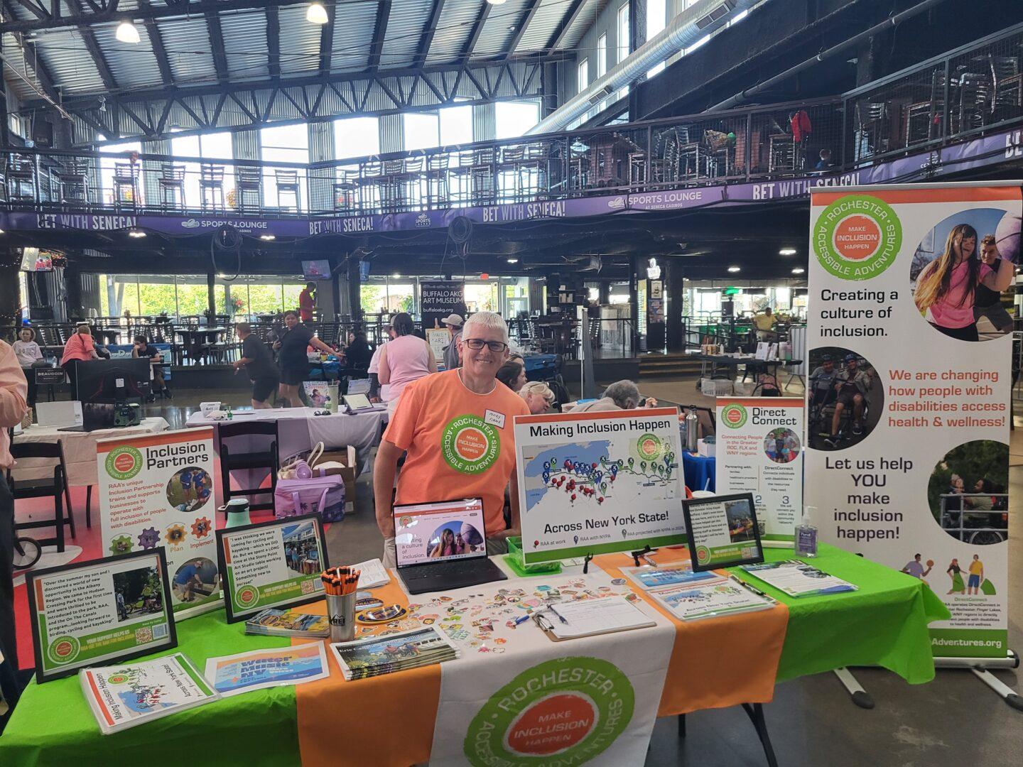 A woman with an RAA shirt on stands at RAA's booth surrounded by informational displays and colorful banners inside the spacious, sunlit Canalside venue.
