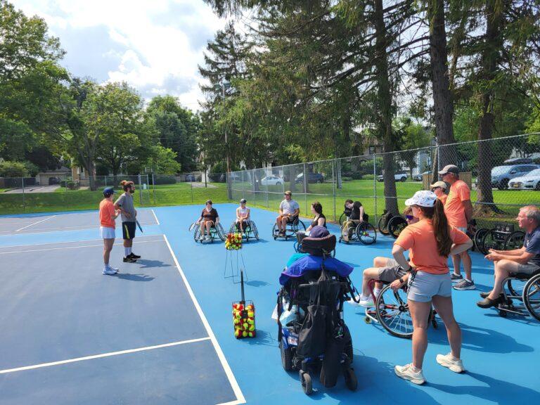 A group of tennis clinic participants in a semi circle listen as the coaches kick off the red ball clinic at an outdoor tennis court painted blue with trees lining the sides