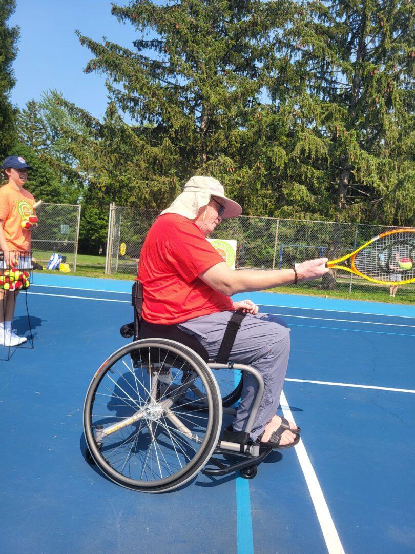 A player wearing a sun hat and a red shirt swings a tennis racket on a blue outdoor court under clear blue skies.