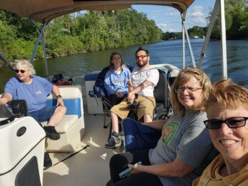 Group of five people smiling for a selfie on board a pontoon boat on a wide river lined by summer green trees
