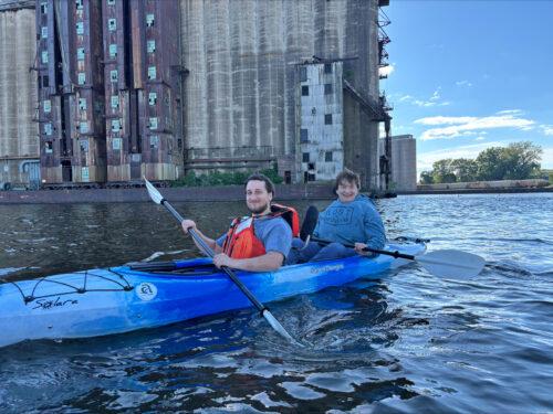 Two people in a blue tandem kayak enjoy calm waters at Elevator Alley Kayak with tall silos behind them