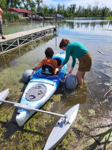 A person up to her knees in water is helping launch a person in a blue adaptive kayak with outriggers along a boardwalk and large pond like entry to the Canal