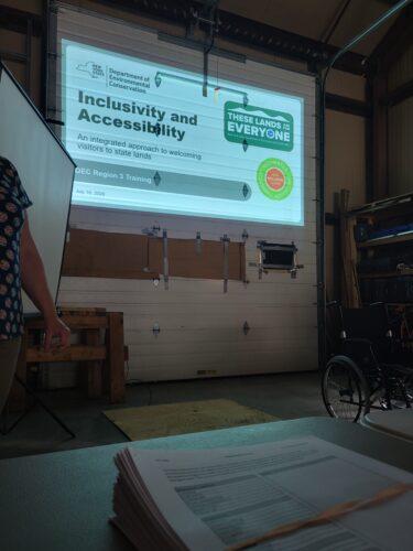 A presentation slide about inclusivity and accessibility for welcoming visitors to state lands, projected on a screen in a workshop classroom setting. Starting in the classroom — grounding the day in what it really means to welcome every visitor to New York's state lands.