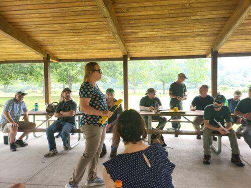 An instructor stands amidst a group of staff seated a tables under a pavilion. Workshop participants gathered around tables under an open pavilion, meeting between field sessions.
