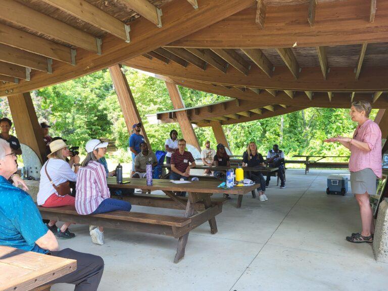 A group of people seated at tables under a pavilion listen to a woman standing at the front