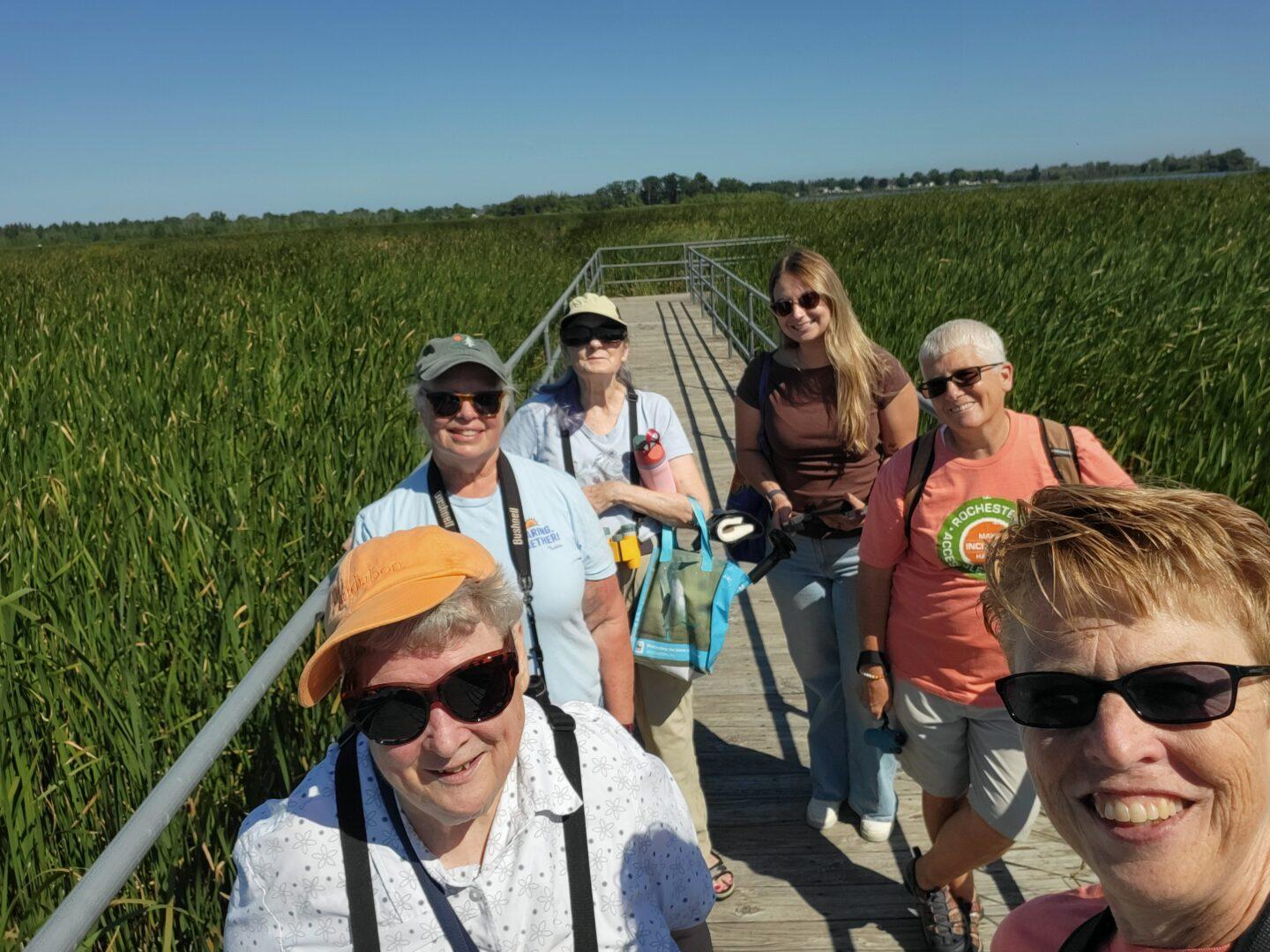 A group of six women on a boardwalk with a large expanse of high reeds and water behind them; they all have birding equipment like binoculars around their necks.