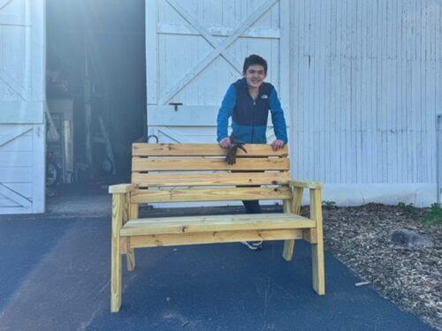 A youth stands behind a newly built wooden bench with arm rests on a paved area in front of a garage
