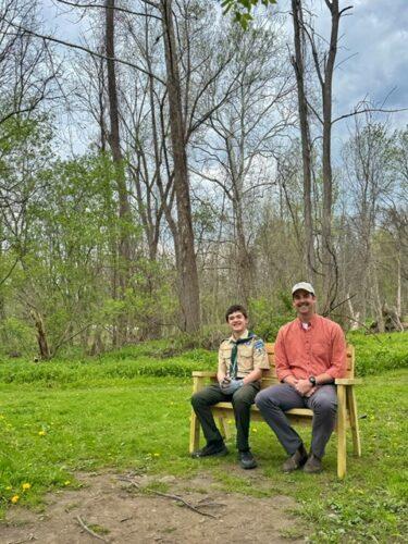 A youth in a scout uniform sits on a newly installed wooden bench in a wooded area with fresh spring leaves on the trees behind them.