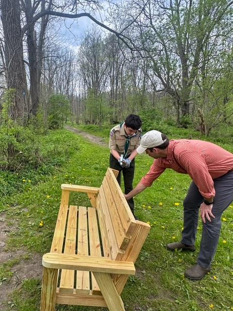 A scout in uniform drills a support on a wooden bench while a man supervises the placement in a wooded area along a path.