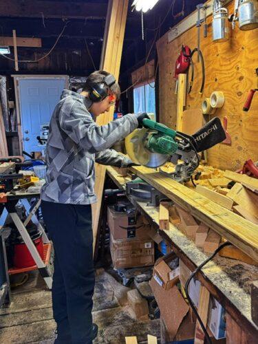 A teen operates a table saw in a garage, wearing headphones and protective gloves.