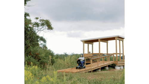 A ramped wooden viewing platform overlooking a wide expanse of water and reeds, a woman on a scooter is coming down the ramp
