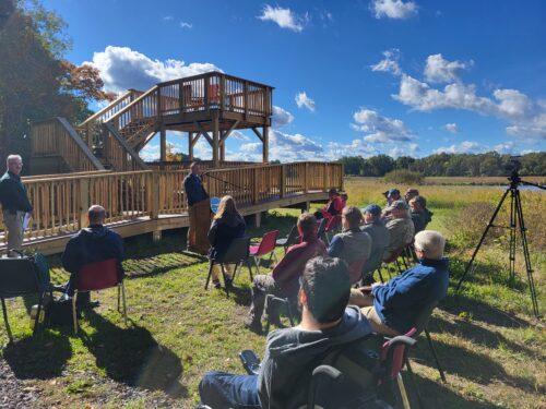 A small group of people are seated in chairs listening to speaker standing at a podium in front of a two story wildlife viewing platform on a bright sunny day with blue sky and puffy white clouds.