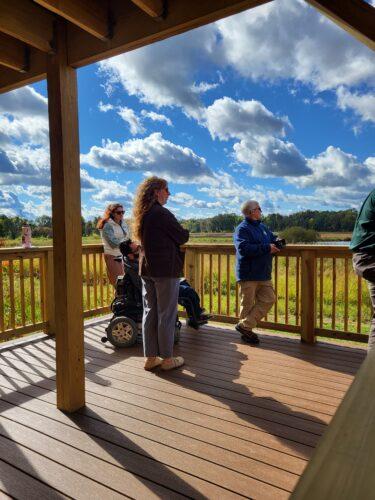 Four people, three standing and one in a power wheelchair, are on a viewing platform looking out across a wide expanse of a nature wildlife area with prairie, water, trees and big blue sky with great puffy white clouds.