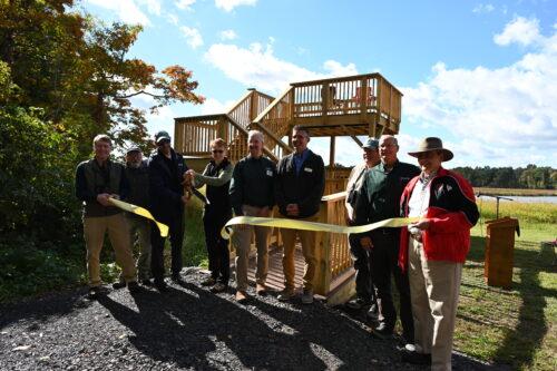 A group of seven people, including RAA's Executive Director, pose to cut a yellow ribbon in front of a ramped viewing platform that has two levels.