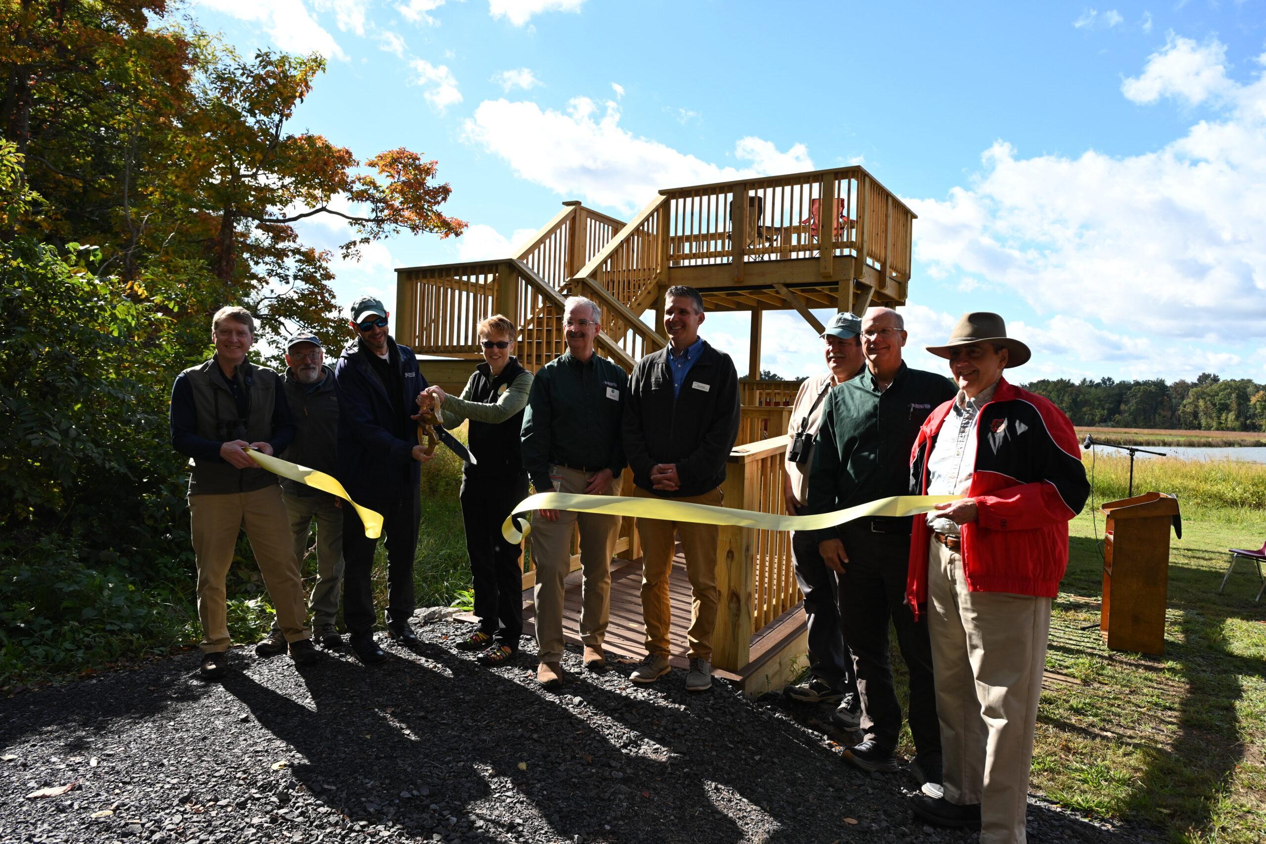 A group of seven people, including RAA's Executive Director, pose to cut a yellow ribbon in front of a ramped viewing platform that has two levels.