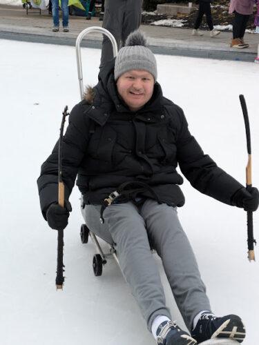 A man on a hockey sled posed with a hockey stick in each  hand on either side of the sled as he smiles on an outdoor ice rink