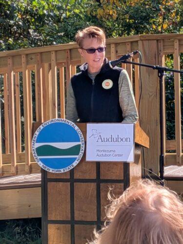 A woman with short red hair and sunglasses with a black vest with RAA's logo is at a podium with the logos of Audubon and NYS DEC; a railing is in the background.